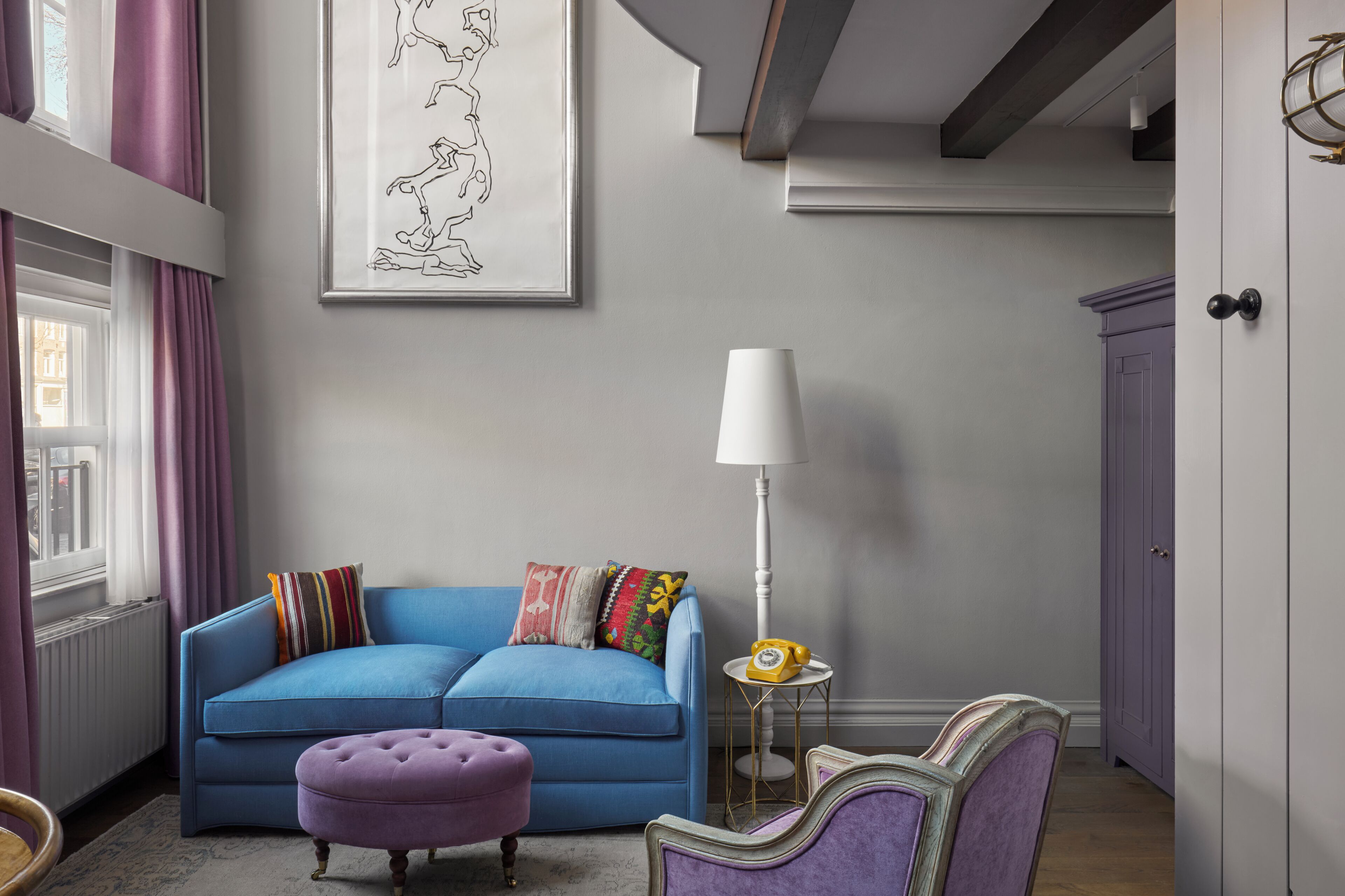 Una sala de estar con un sofá azul, un reposapiés morado, una lámpara blanca y un mueble morado.