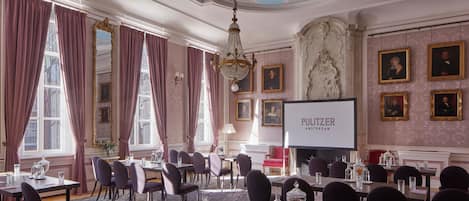 A conference room with tables, chairs, a projector screen, and framed portraits on the walls.
