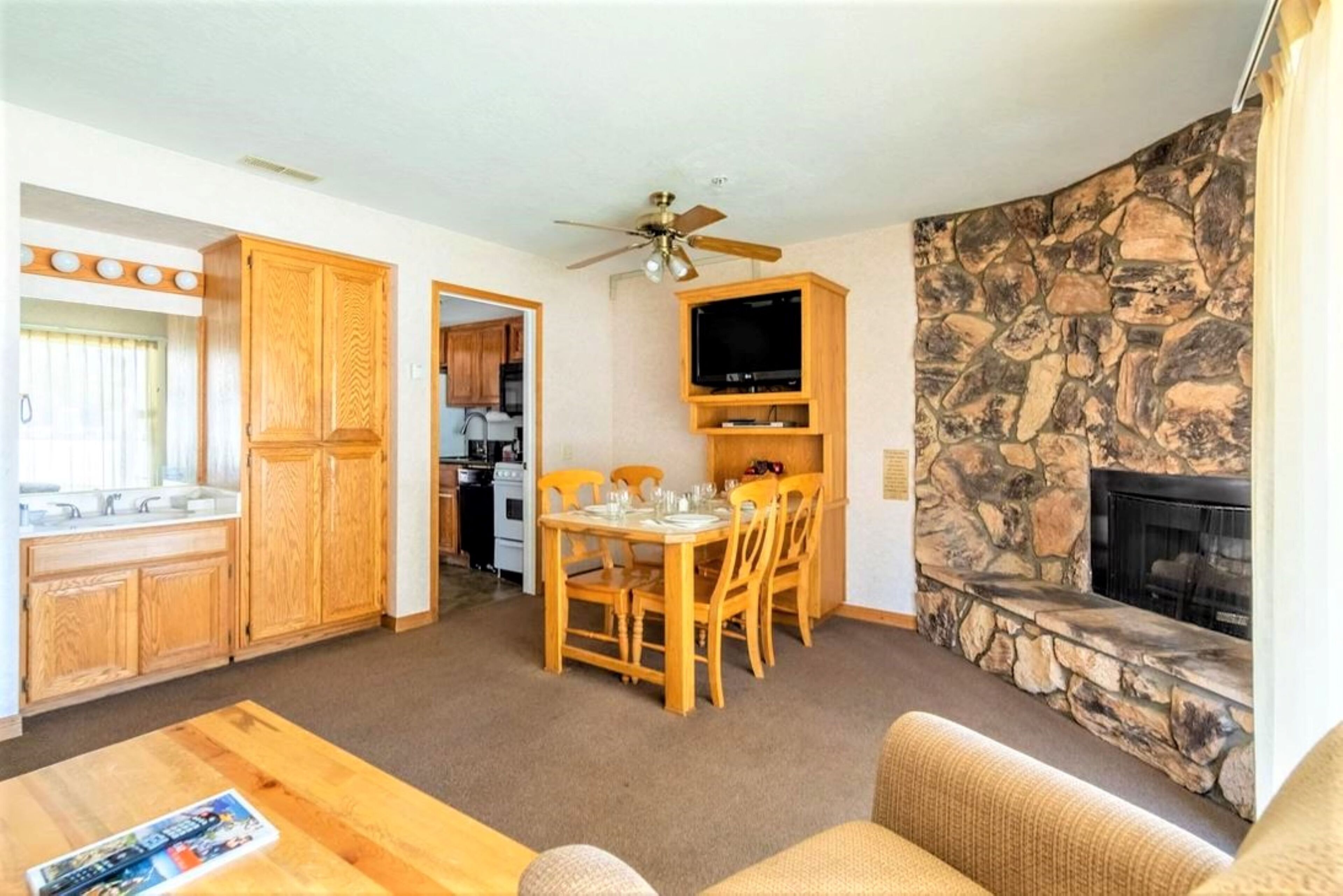 A cozy living room with a stone fireplace, wooden dining table, and a television mounted above it.