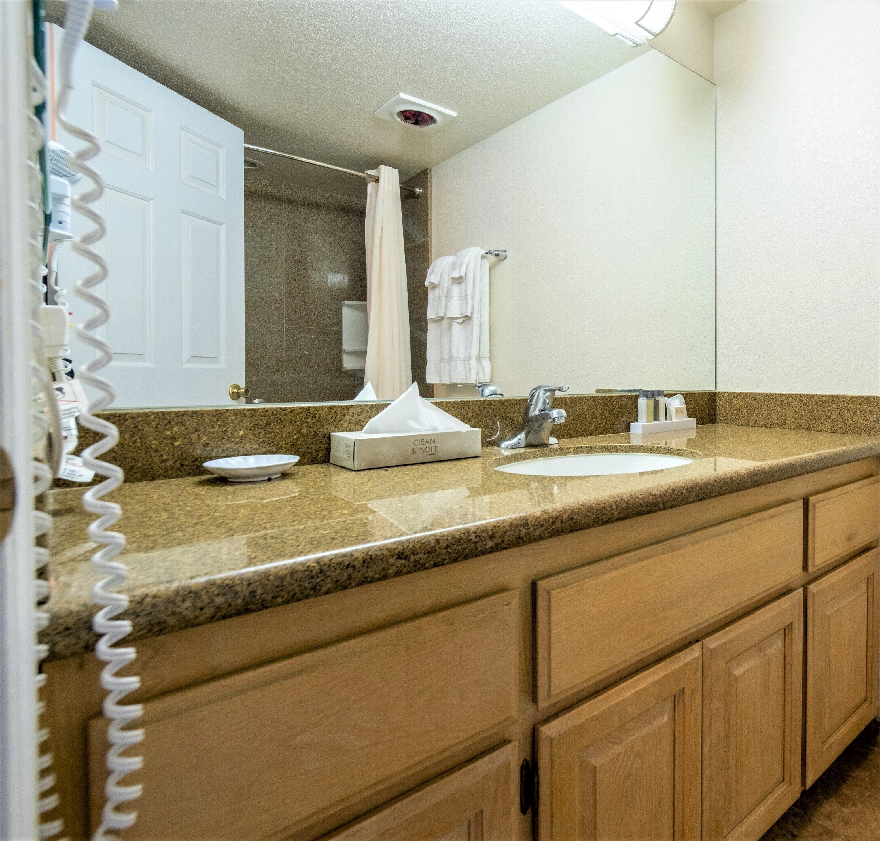 A hotel bathroom with a granite countertop, a sink, a mirror, and a shower area.