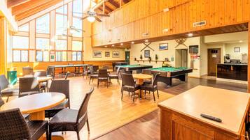A spacious indoor area with wooden walls and ceiling, featuring a pool table, billiards, and multiple seating areas with tables and chairs.
