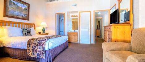 A bedroom with a bed, bedside table, lamp, and a large mirror. There is a second floor balcony visible.