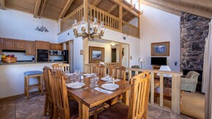 A spacious dining area with wooden furniture and a chandelier, a kitchen with modern appliances, and a stone fireplace. - Lagonita Lodge (Big Bear Lake)
