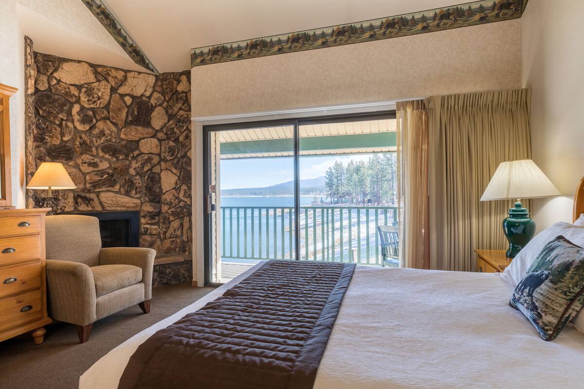 Een slaapkamer met een stenen muur, een bed met een bruin hoofdeinde, een houten dressoir, een stoel en uitzicht op de omgeving via een schuifdeur.