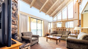 A spacious living room with a stone fireplace, large windows, and a high wooden ceiling. - Lagonita Lodge (Big Bear Lake)