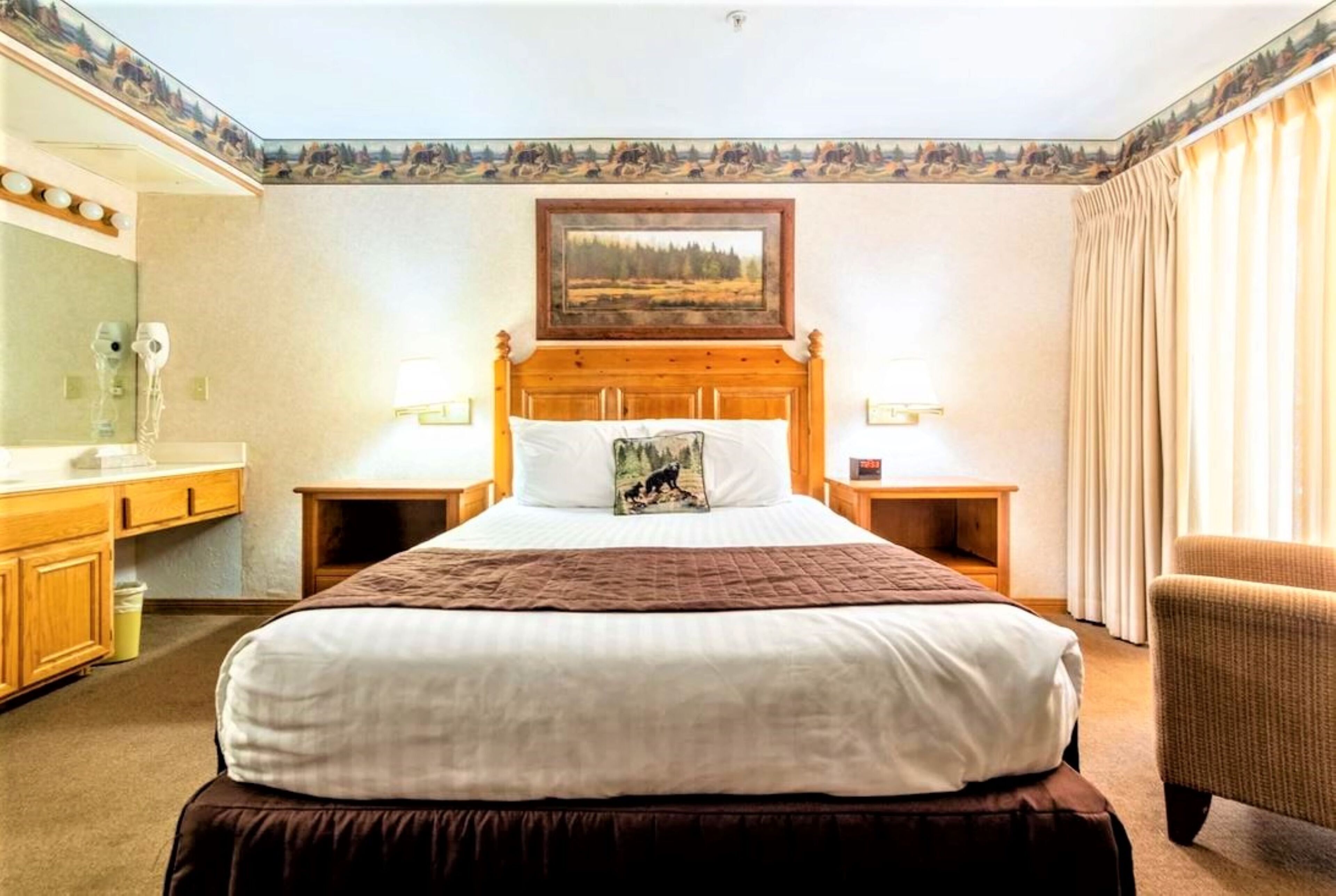 Ein Hotelzimmer mit einem großen Bett, einem hölzernen Kopfteil, einem Bild über dem Bett, zwei Nachttischlampen, einem Sessel, einem kleinen Tisch, einem Spiegel und einem Fenster mit Vorhängen.