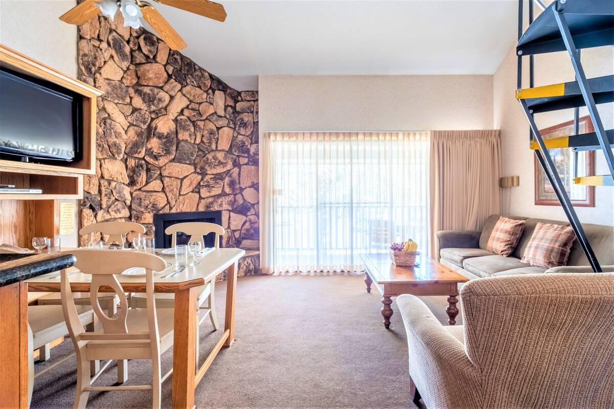 Een woonkamer met een stenen muur, een televisie, een eettafel met stoelen, een bank en een trap.