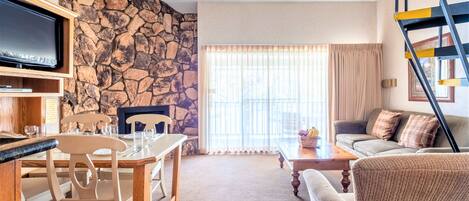 Una sala de estar con pared de piedra, televisor, mesa de comedor con sillas, sofá y una escalera.