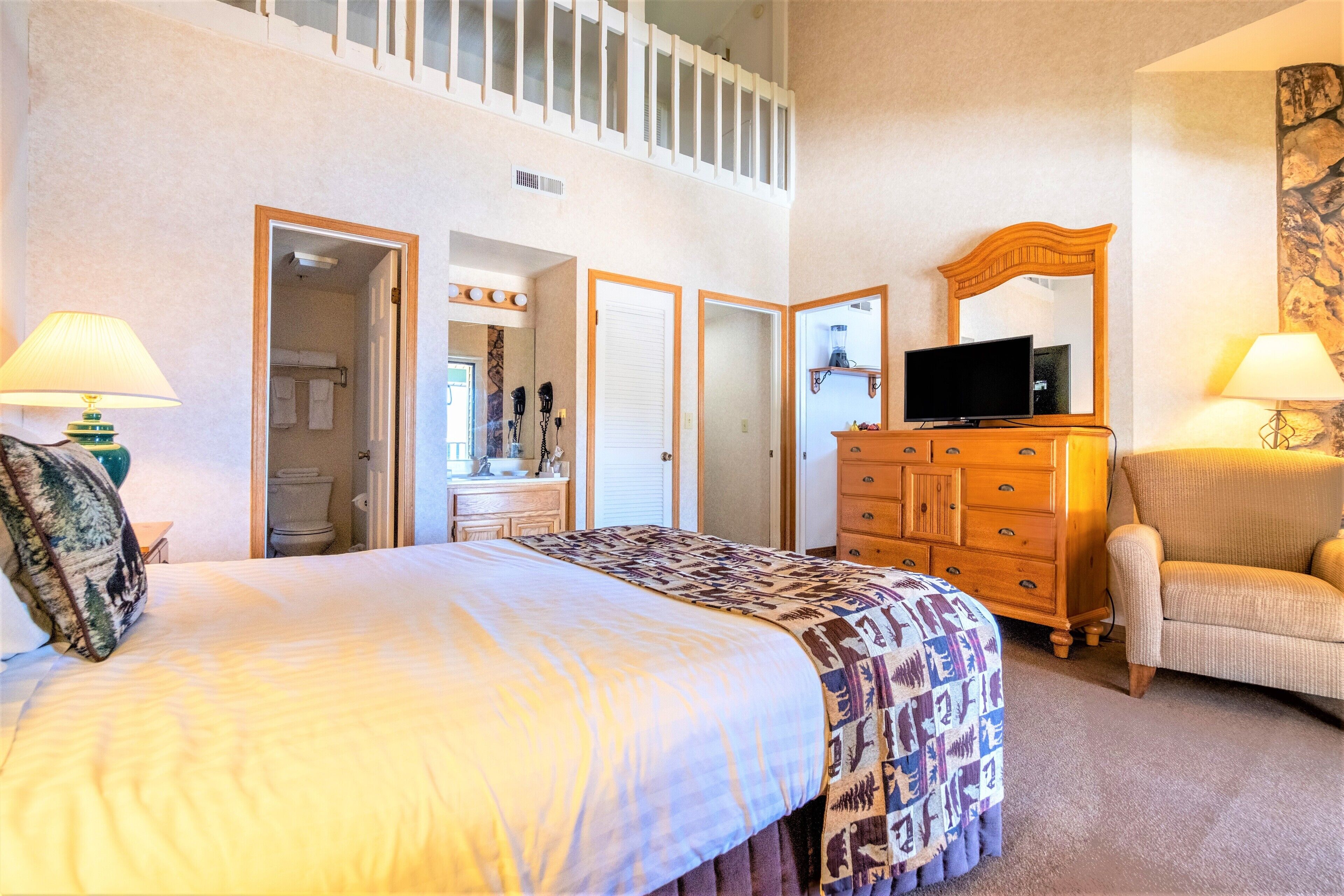 Une chambre d’hôtel avec un grand lit, une commode, une télévision et une chaise. On y trouve des portes donnant accès à une salle de bain et à un placard.