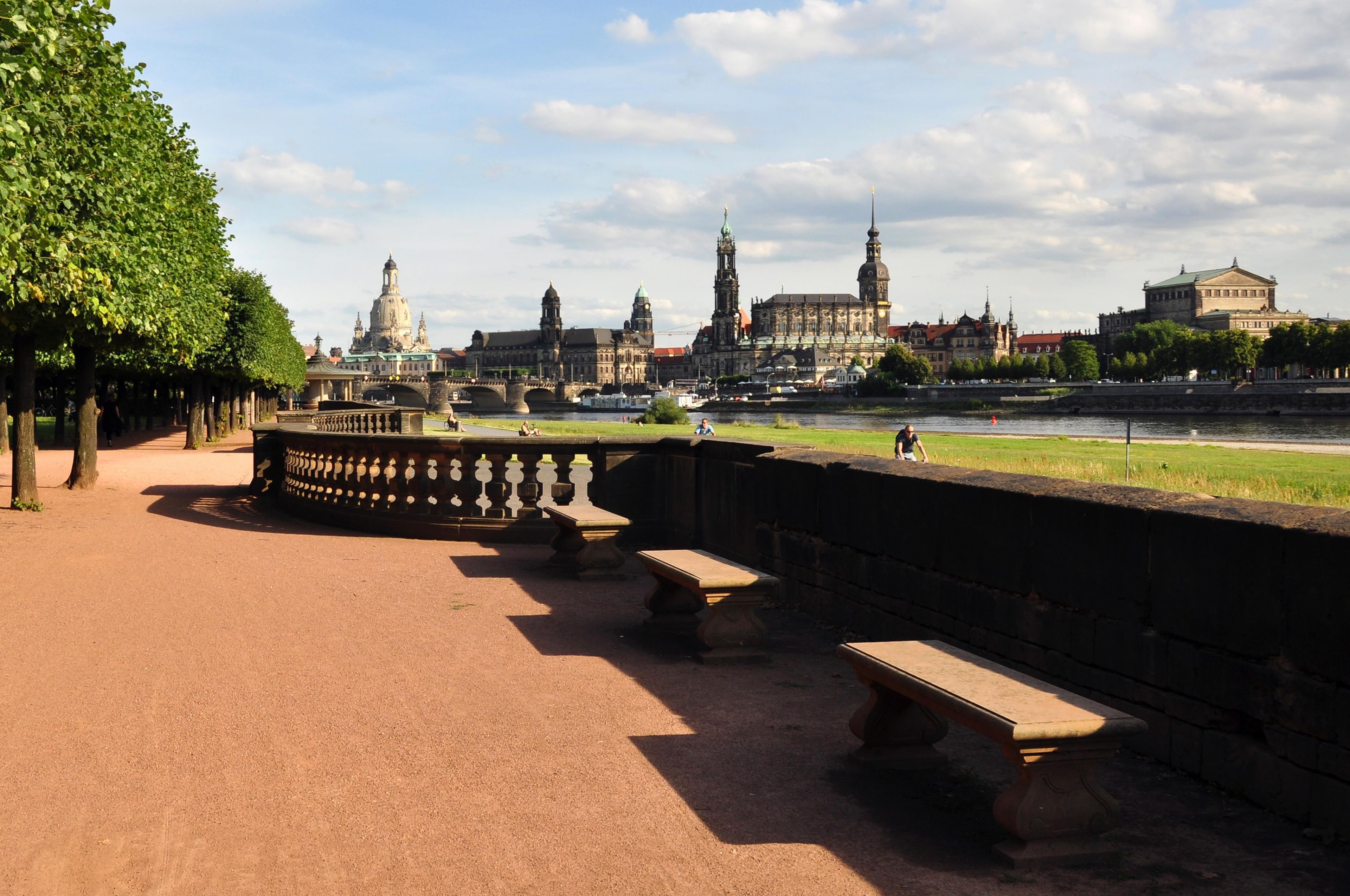 Une promenade au bord de la rivière, agrémentée de bancs et d’une balustrade en pierre, qui mène à un ensemble de bâtiments historiques comprenant plusieurs tours et coupoles.