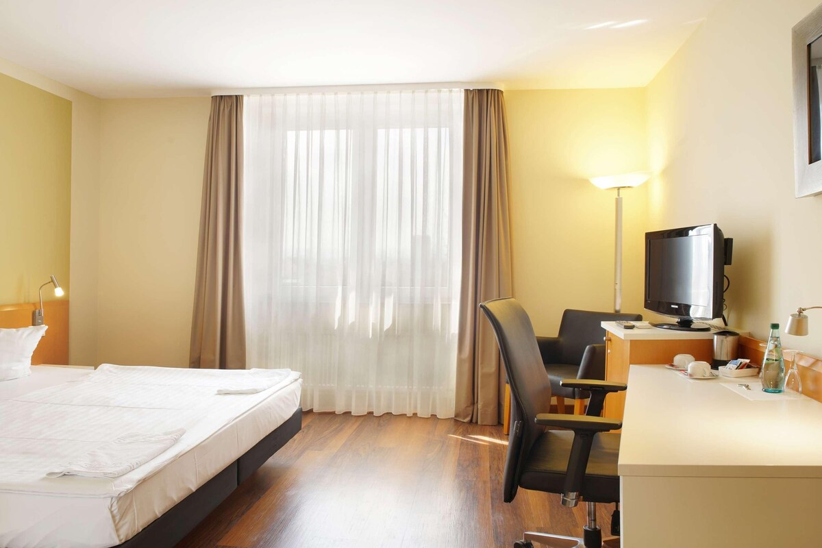 A hotel room with a bed, a desk with a computer, a chair, a television, and a window with curtains.