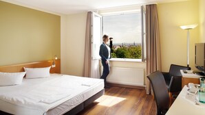 A hotel room with a bed, a desk, a chair, a window with a view of a cityscape, and a lamp.