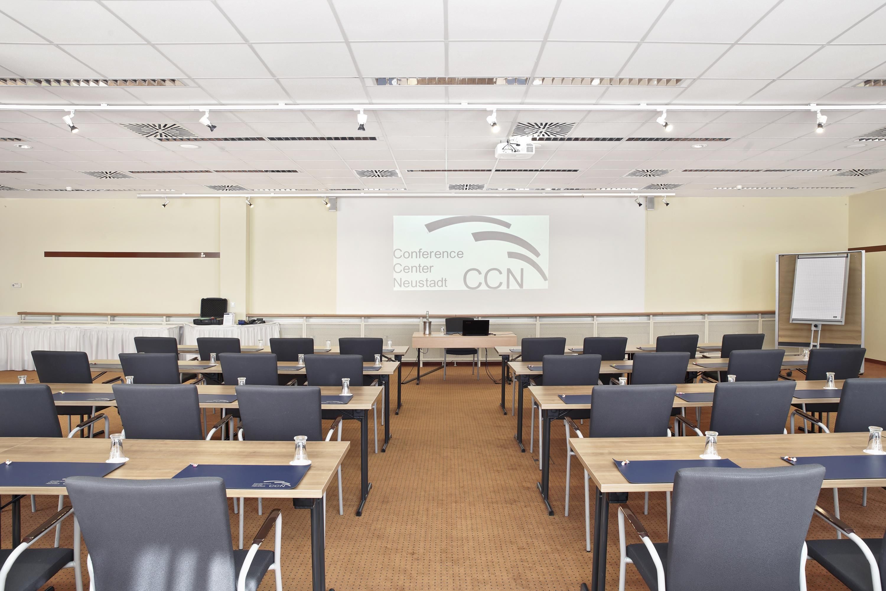 A conference room with rows of tables and chairs, a projection screen displaying 'Conference Center Neustadt CCN', and a whiteboard on the right.