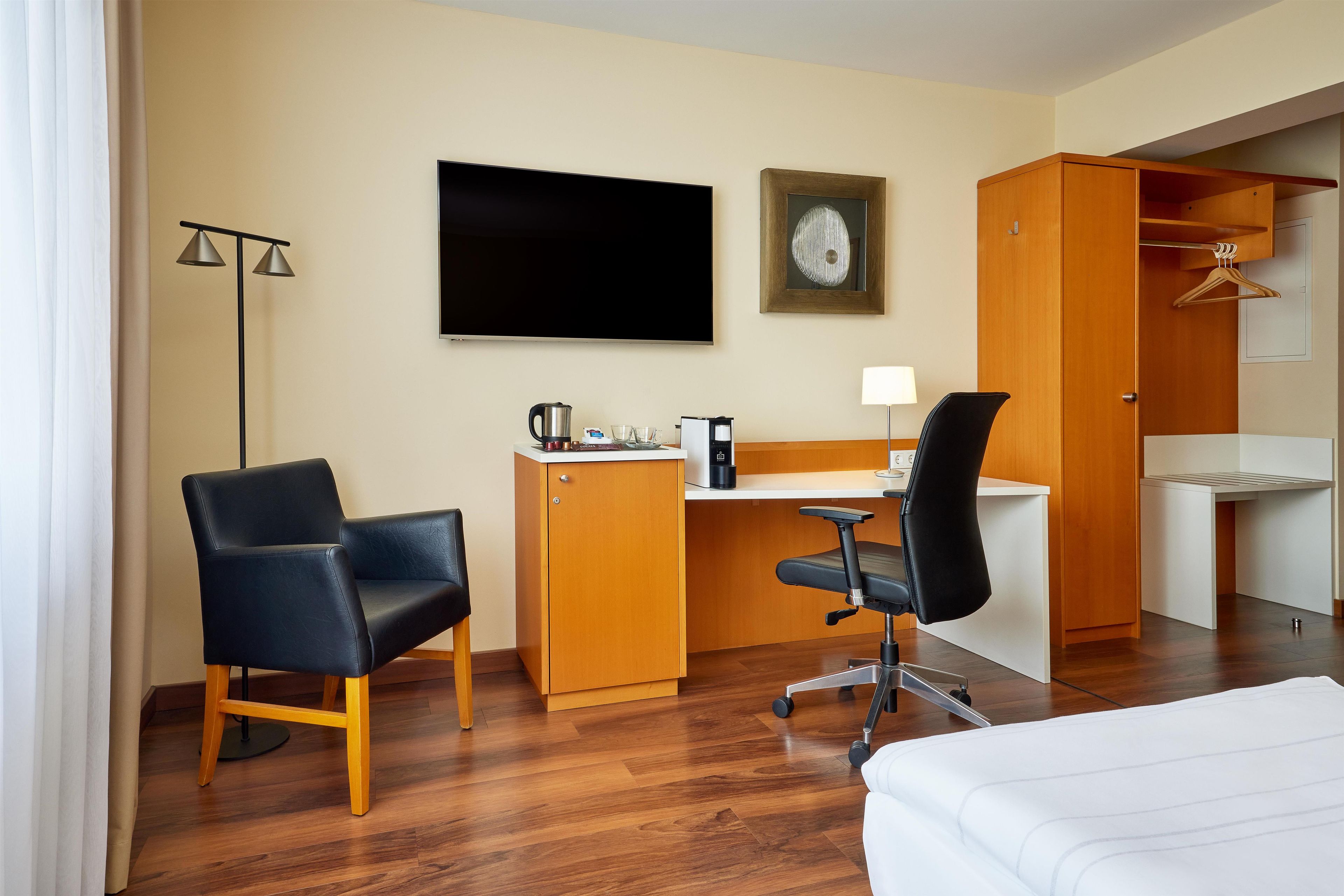 A hotel room with a bed, a desk with a chair, a television, a wardrobe, and a floor lamp.