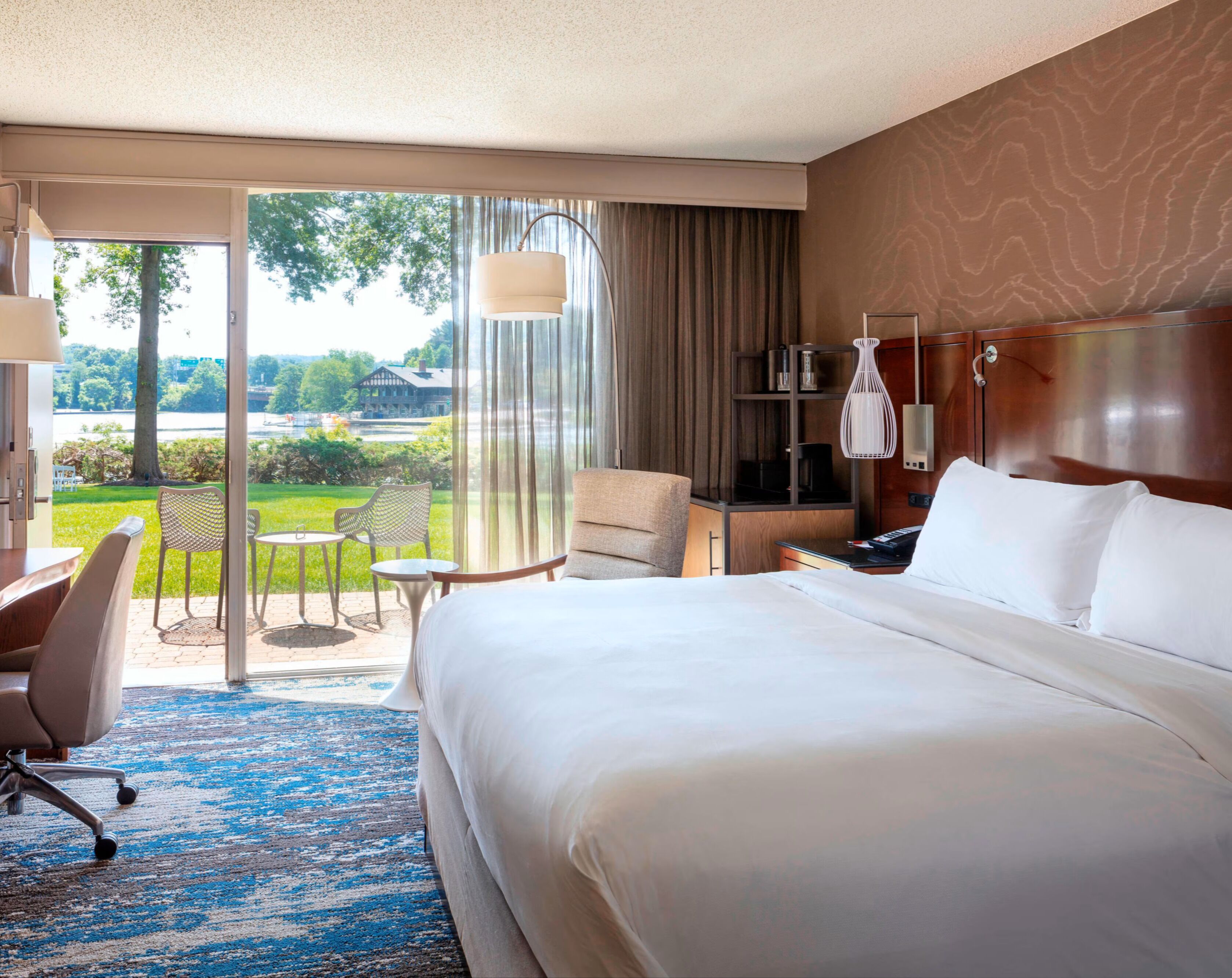 Habitación de hotel con una cama grande, un escritorio, una silla, una mesita y vistas a un jardín y un edificio a través de una puerta de cristal.