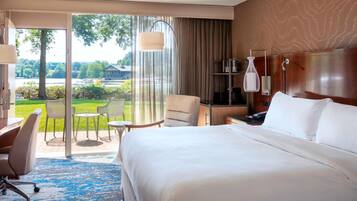 Habitación de hotel con una cama grande, un escritorio, una silla, una mesita y vistas a un jardín y un edificio a través de una puerta de cristal.