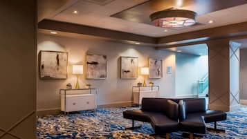 Eine Hotelhalle mit blauem, gemustertem Teppich, einer schwarzen Ledersofa, zwei weißen Beistelltischen mit Lampen und abstrakter Wandkunst.
