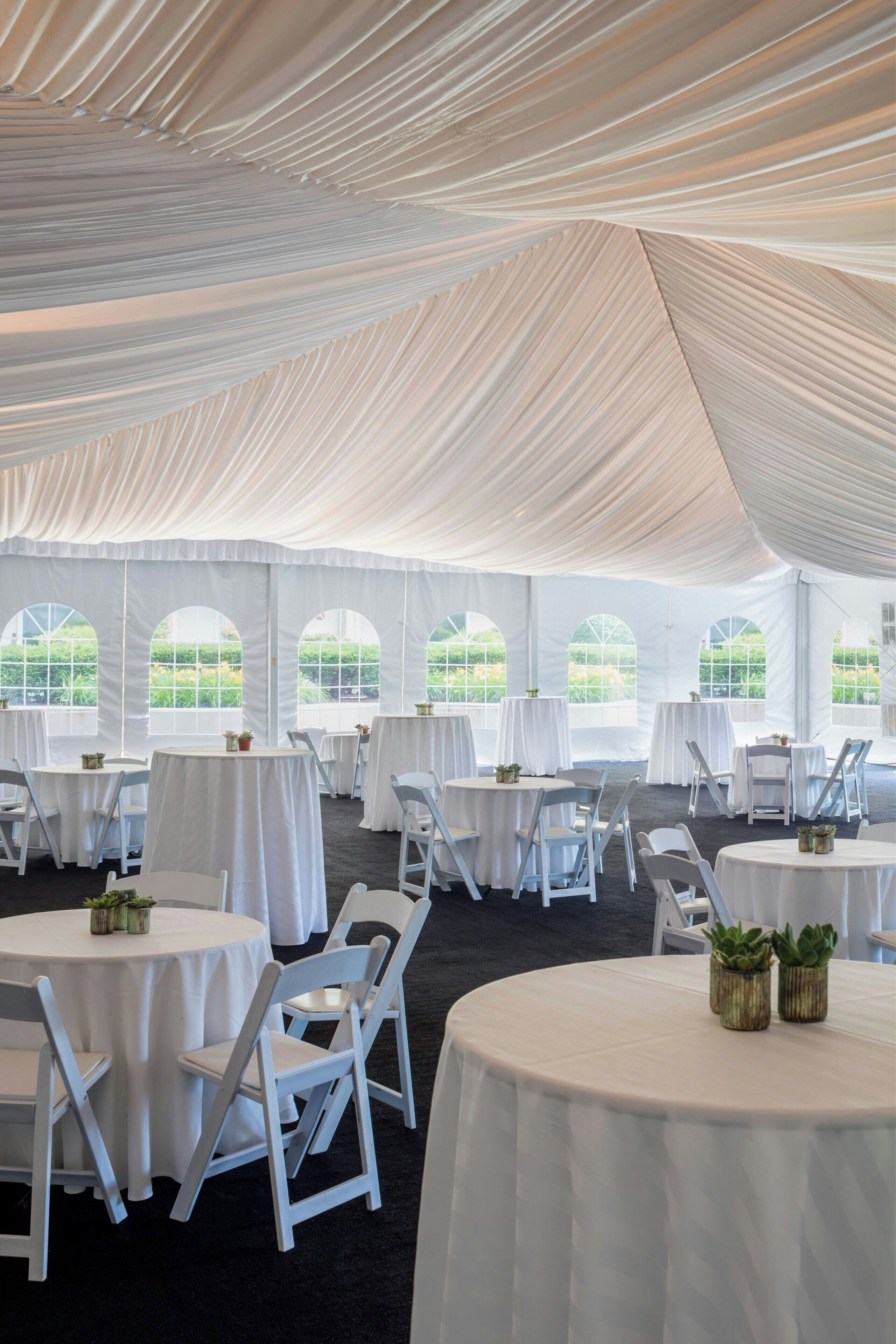 A tented event space with round tables and white chairs, large windows, and a view of greenery outside.