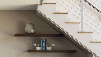 Un salon moderne avec un escalier, un canapé blanc agrémenté de coussins bleus, un fauteuil de bureau gris et des étagères ornées d’objets décoratifs et de livres.