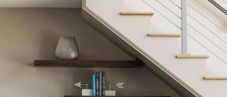 A modern living room with a staircase, a white sofa with blue pillows, a grey office chair, and shelves with decorative items and books.
