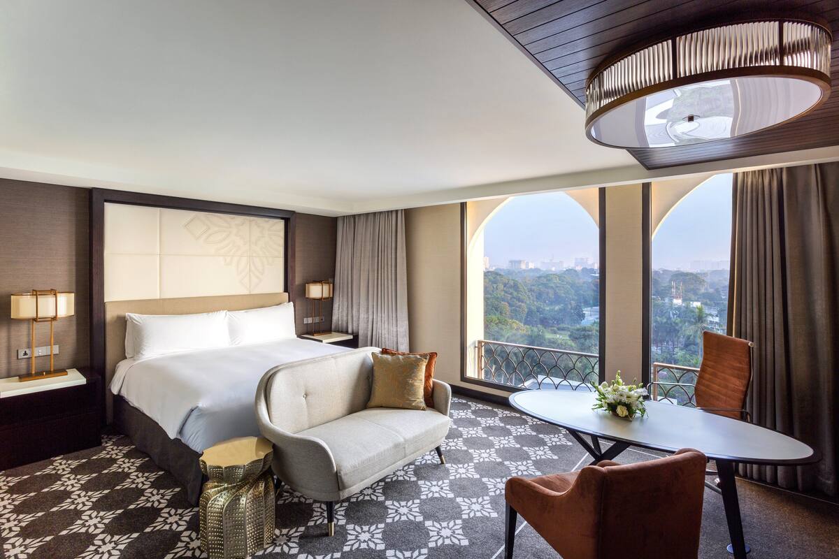 A hotel room with a large bed, a sofa, a dining table with chairs, and a balcony with a view.