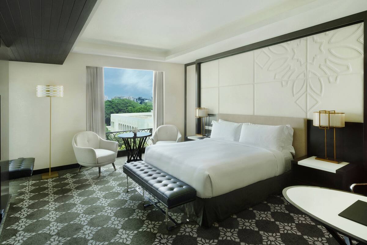 A hotel room with a large bed, a flat-screen TV, a seating area with a table and chairs, and a view of greenery outside.