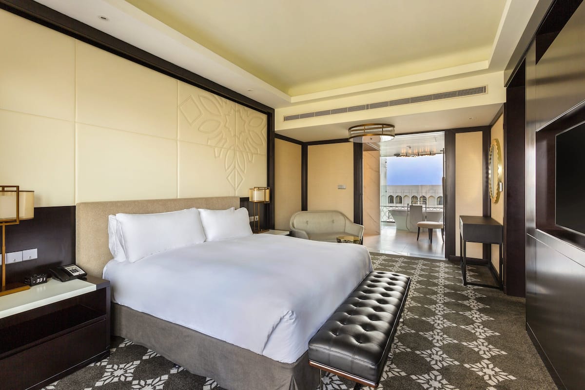 A hotel room with a large bed, a bedside table with a lamp, a chair, a small table, and a TV.