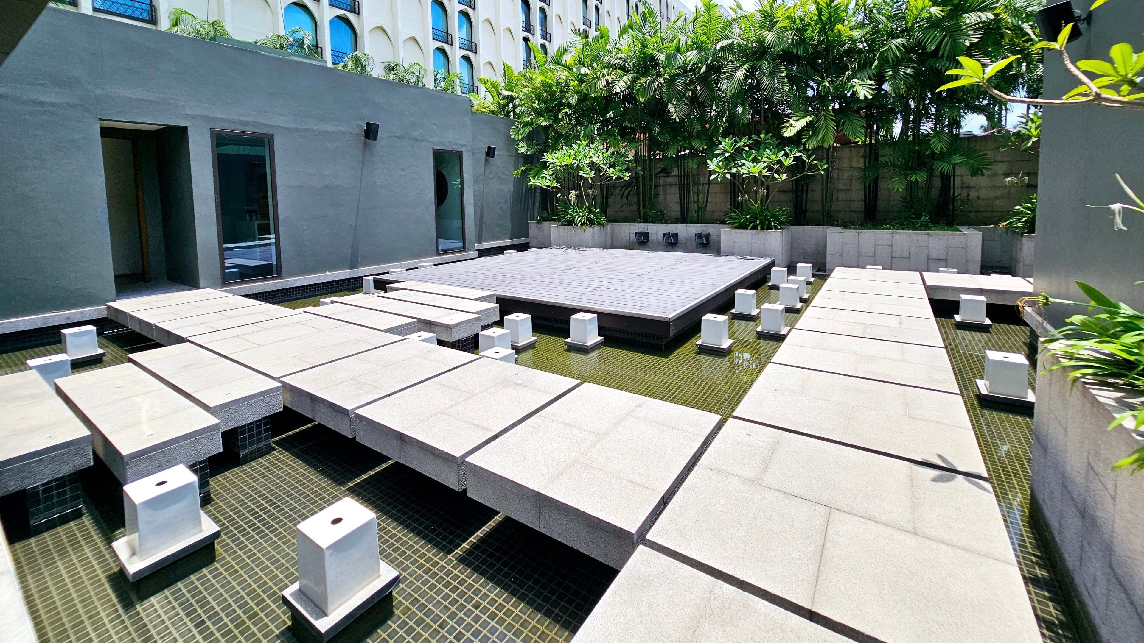 A rooftop terrace with a grid of square tables and chairs, surrounded by greenery and a building with balconies.