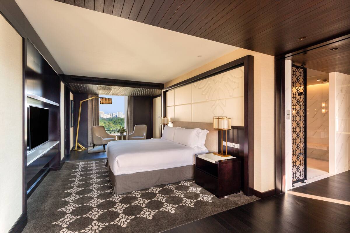 A modern hotel room with a large bed, a flat-screen TV, a seating area with a chair and a table, and a view of the cityscape through the window.