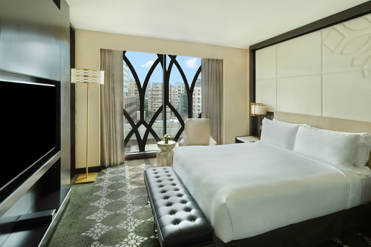 A hotel room with a large bed, a TV, a desk with a lamp, and a view of the city through a window with a geometric design.