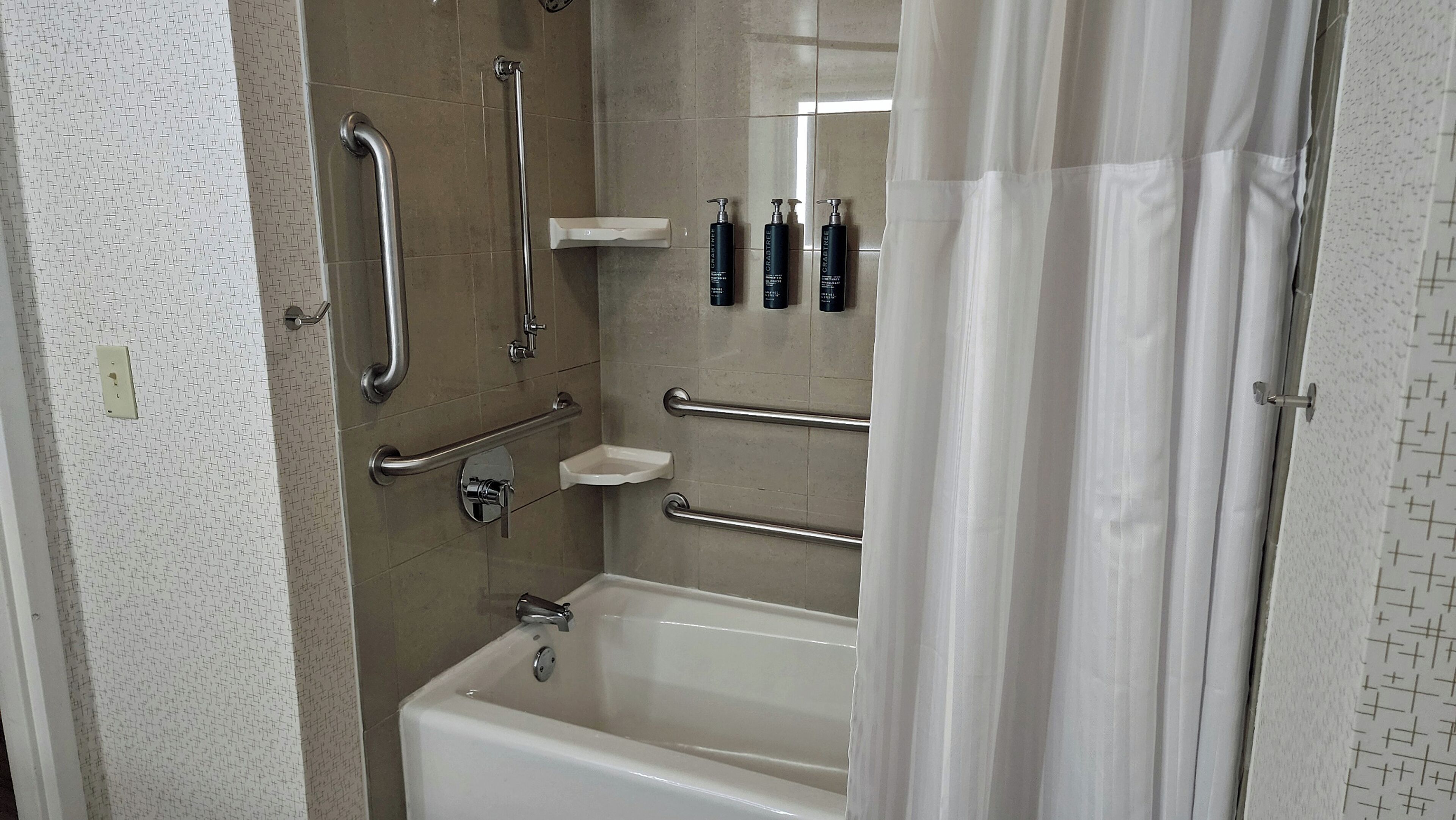 A bathroom with a bathtub, handrails, a shower area, and toiletries on the wall.