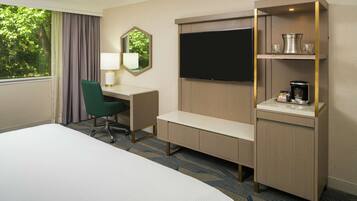 Habitación de hotel con cama, escritorio y silla, televisión, cafetera y estante con vasos.
