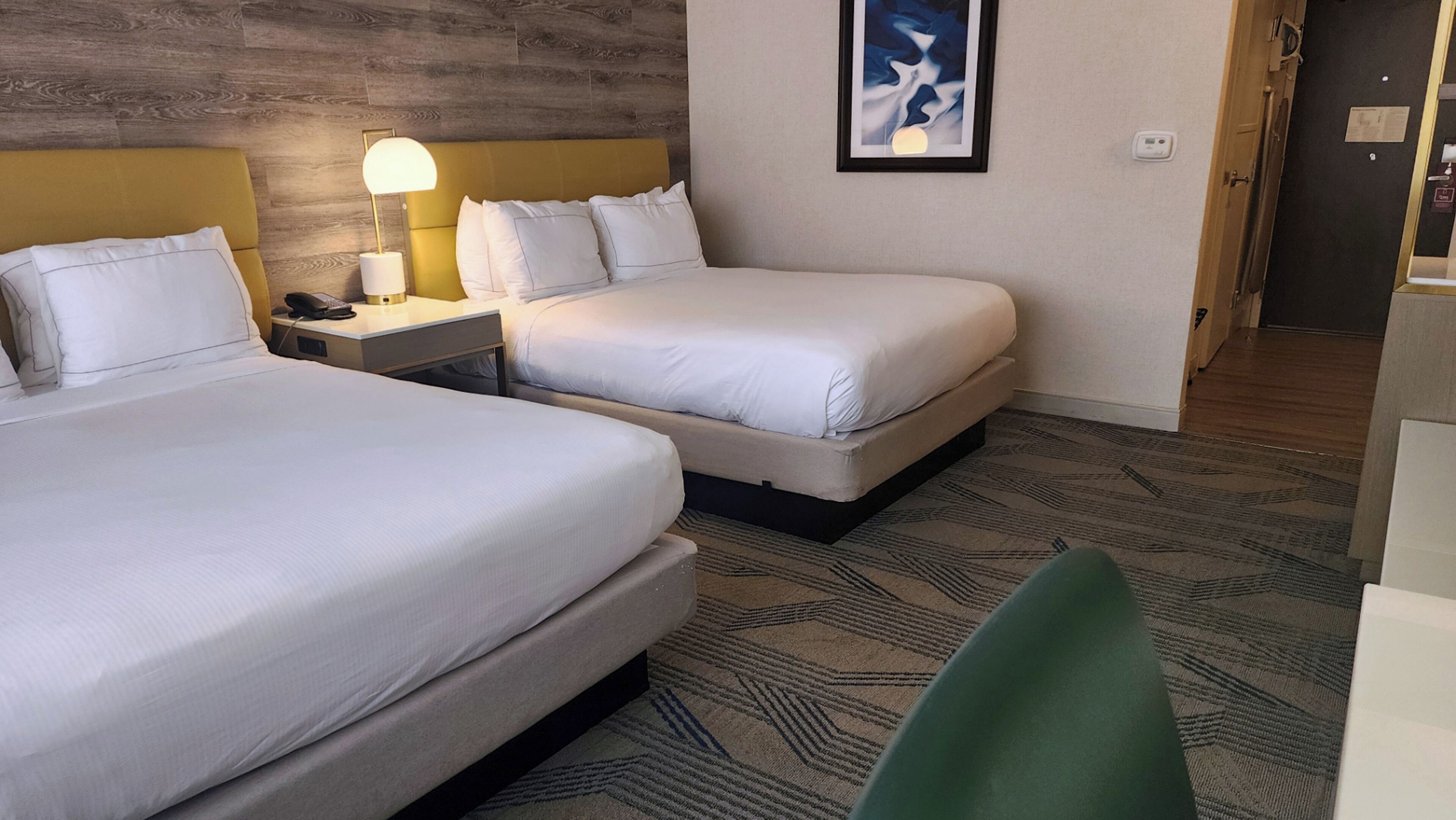 Habitación de hotel con dos camas, una mesita de noche con lámpara, una obra de arte abstracta enmarcada en la pared y una alfombra estampada.