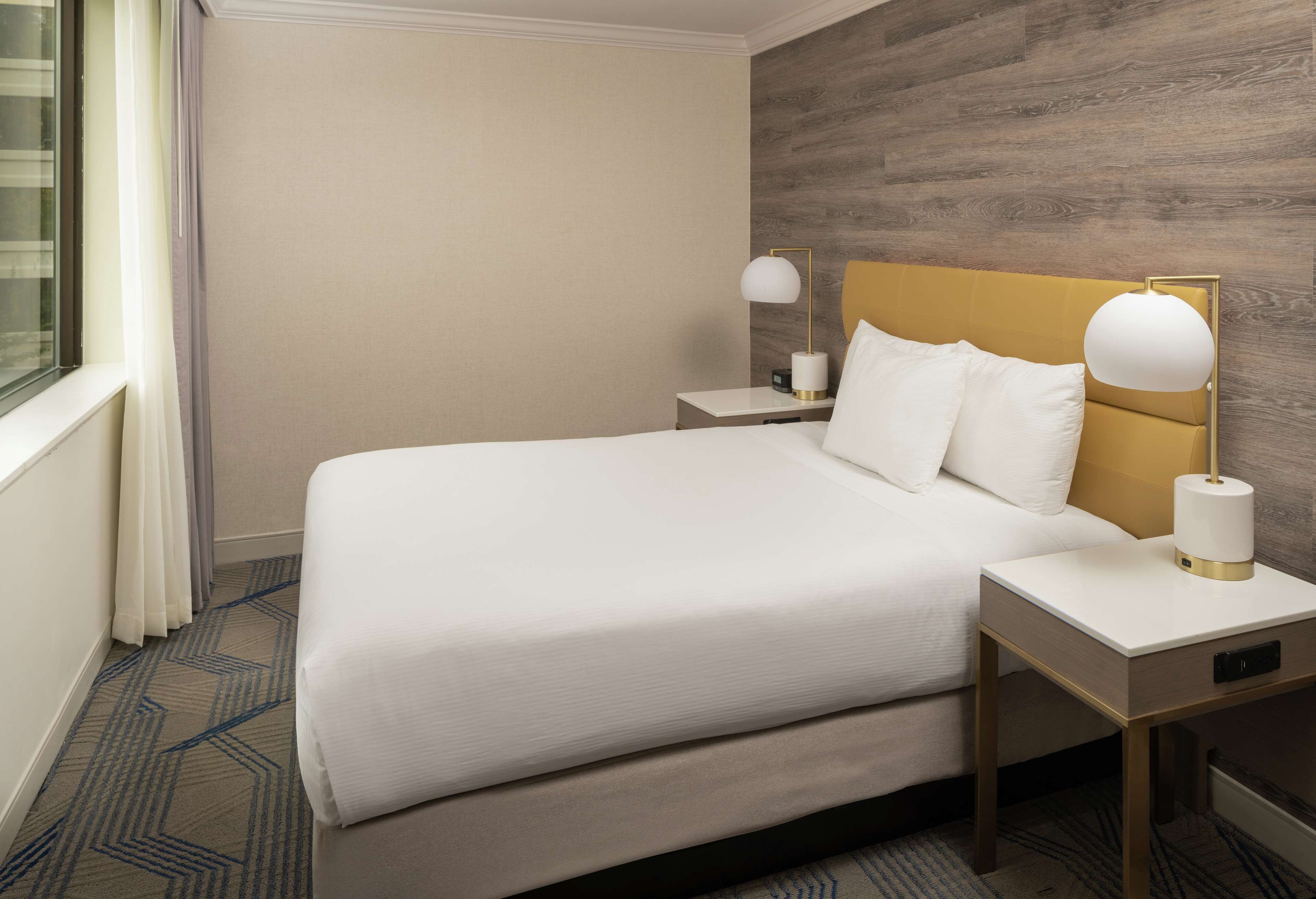 A hotel room with a large bed, a wooden headboard, a nightstand with a lamp, and a window with curtains.