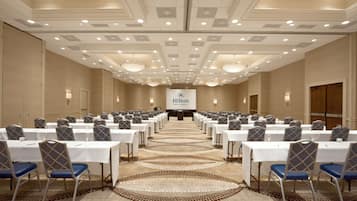 Un salón de conferencias con filas de mesas y sillas, suelo alfombrado e iluminación empotrada en el techo. Se ve una pancarta con el logotipo de Hilton al fondo.