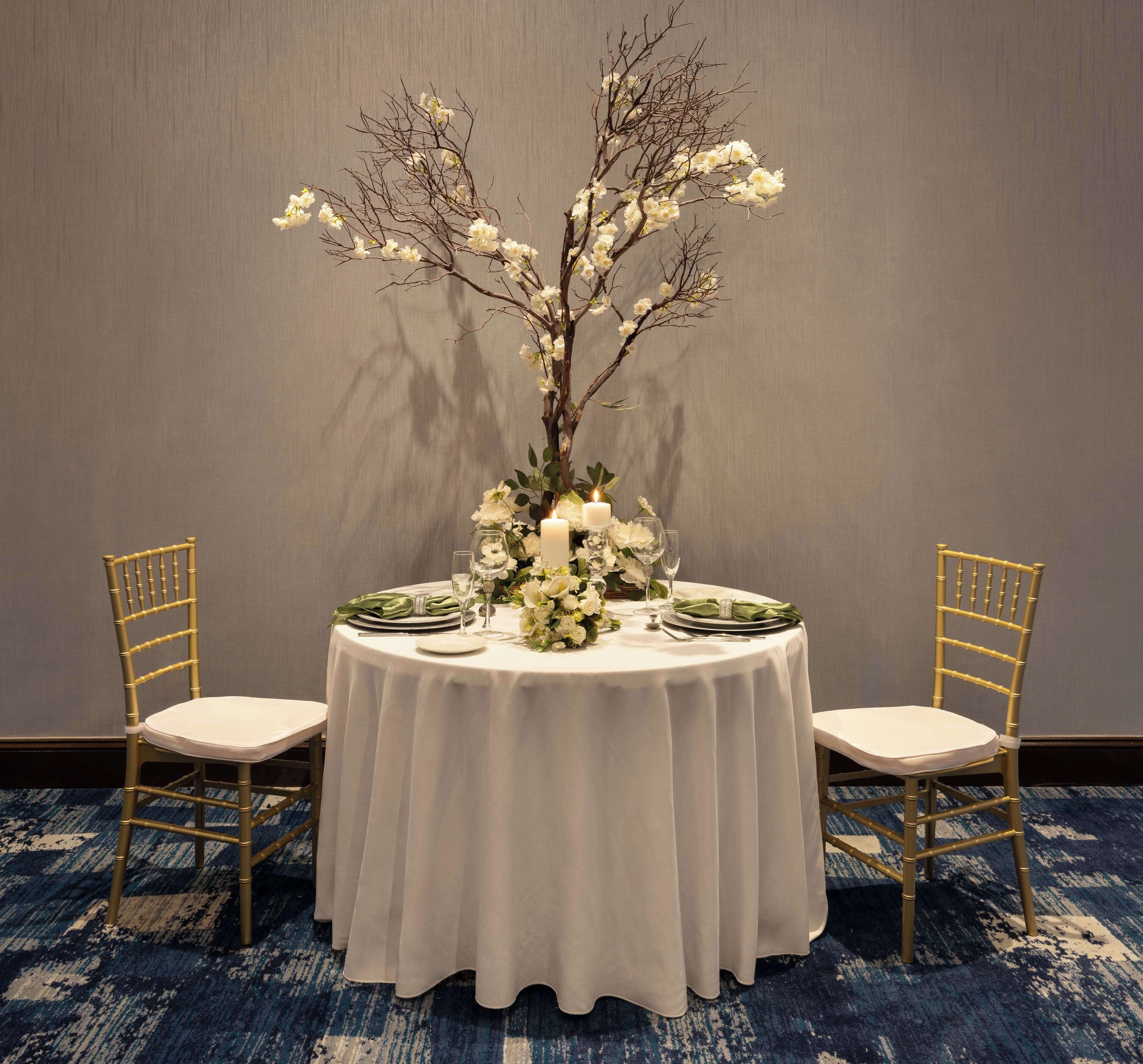 Una mesa redonda con mantel blanco, dos sillas, un centro de mesa floral y una vela encendida.