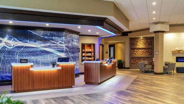Uma área de recepção de hotel moderna, com dois balcões de madeira, uma grande parede de exibição digital e uma área de estar com cadeiras.