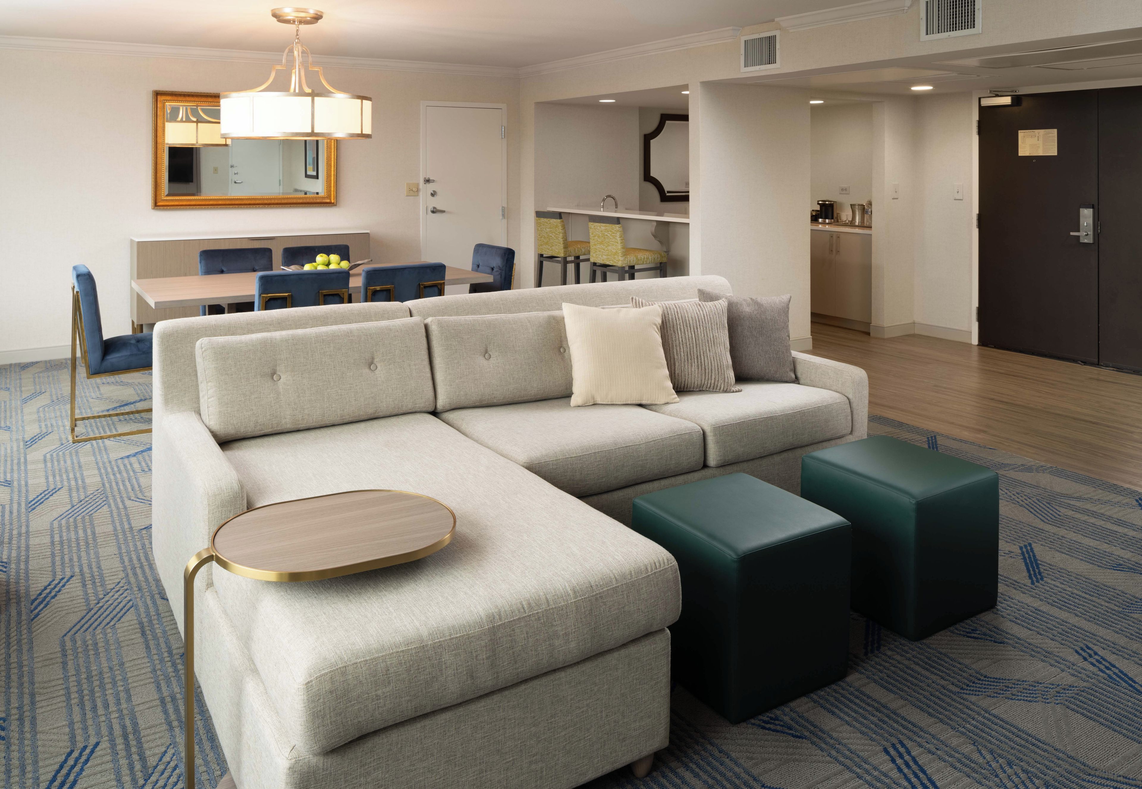 A modern living room with a sectional sofa, a round coffee table, and two ottomans. There is a dining area with a table and chairs in the background.