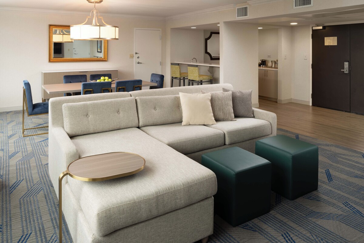 A modern living room with a sectional sofa, a round coffee table, and two ottomans. There is a dining area with a table and chairs in the background.