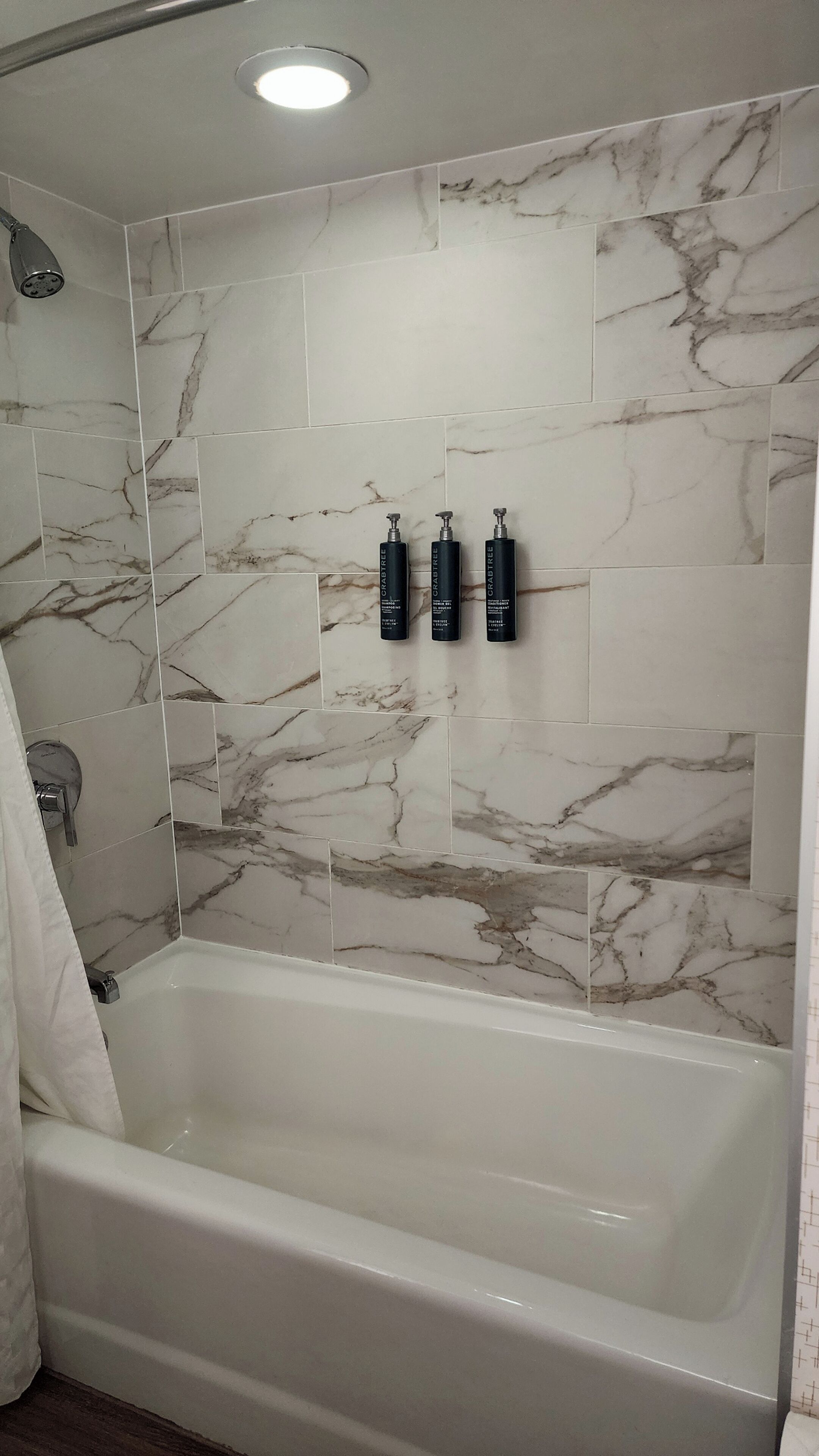 A bathroom with a marble-tiled shower wall, a white bathtub, and three black bottles on the wall.