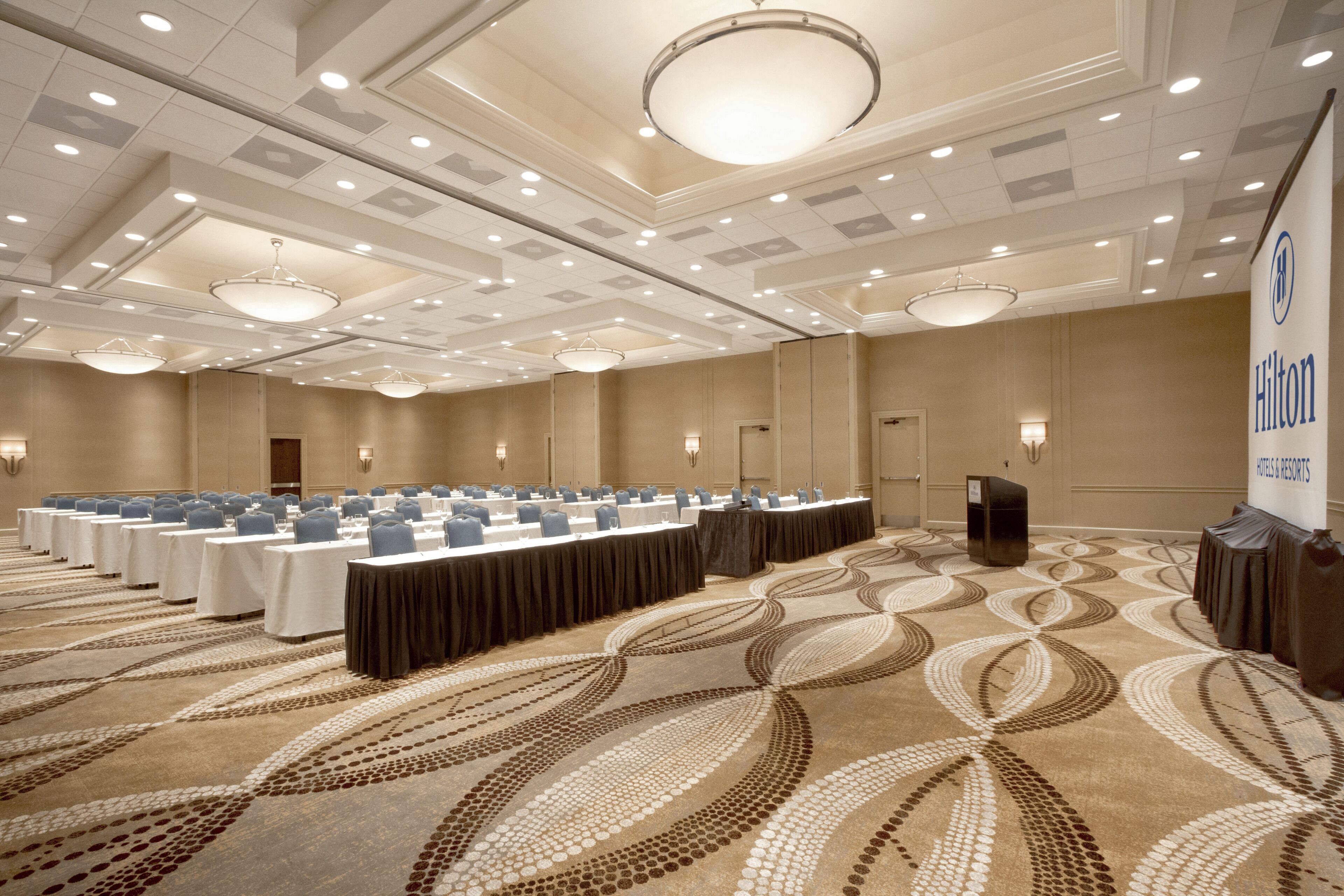 A large conference room with multiple tables, a podium, and a Hilton banner.