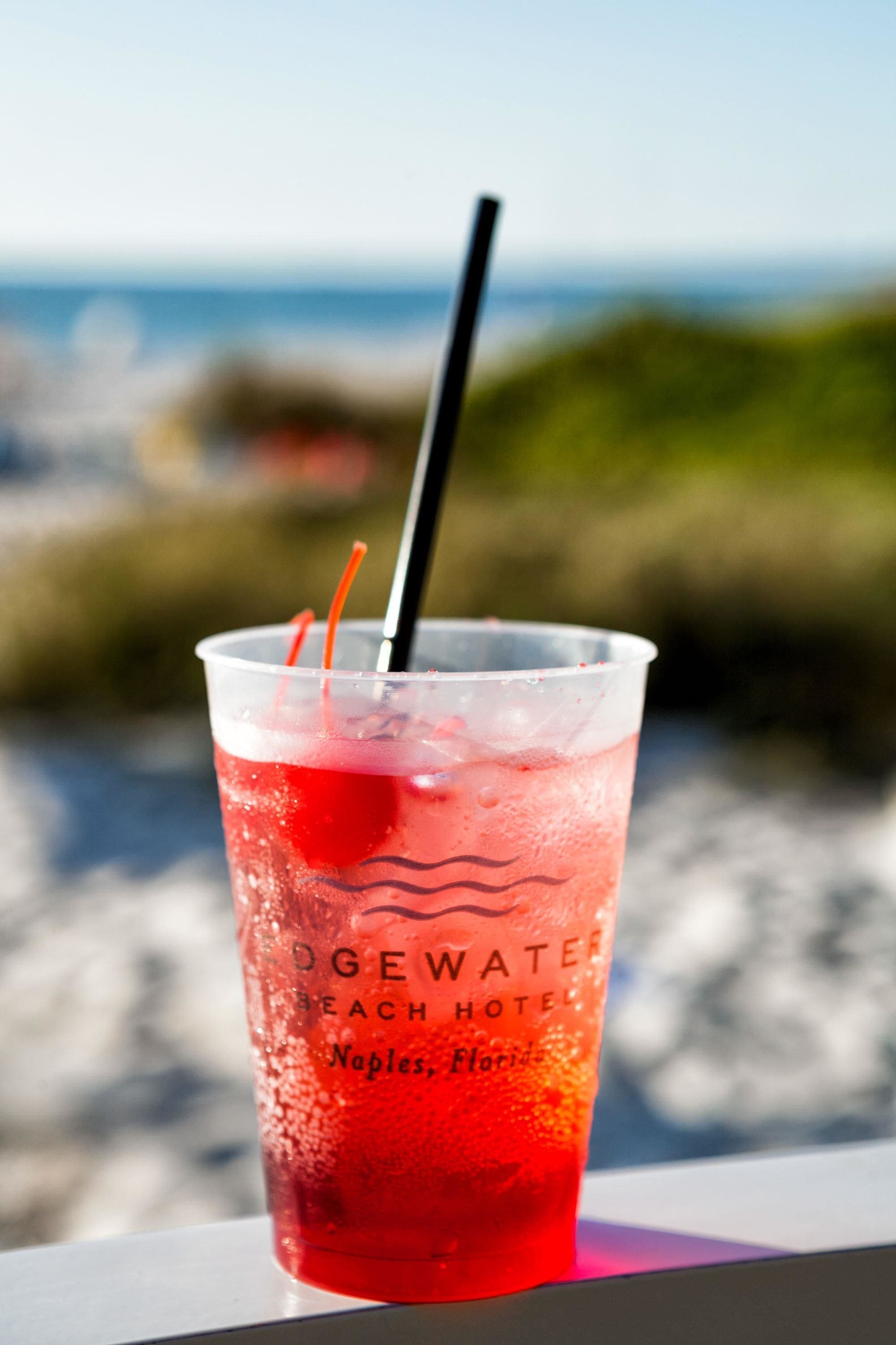 A glass of red beverage with a straw, labeled "Edgewater Beach Hotel Naples, Florida," placed on a ledge with a blurred background of greenery and water.