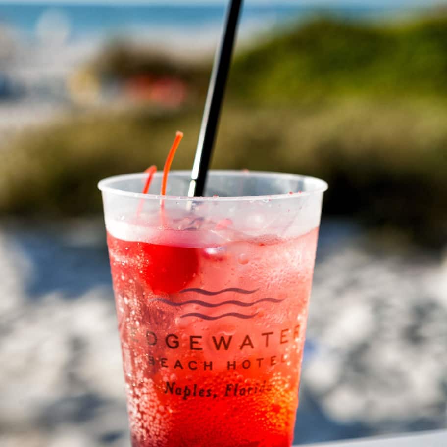 A glass of red beverage with a straw, labeled "Edgewater Beach Hotel Naples, Florida," placed on a ledge with a blurred background of greenery and water.