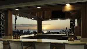 A bar area with a view of the sunset over the ocean, wooden cabinets, and bar stools. - Edgewater Beach Hotel (Naples)