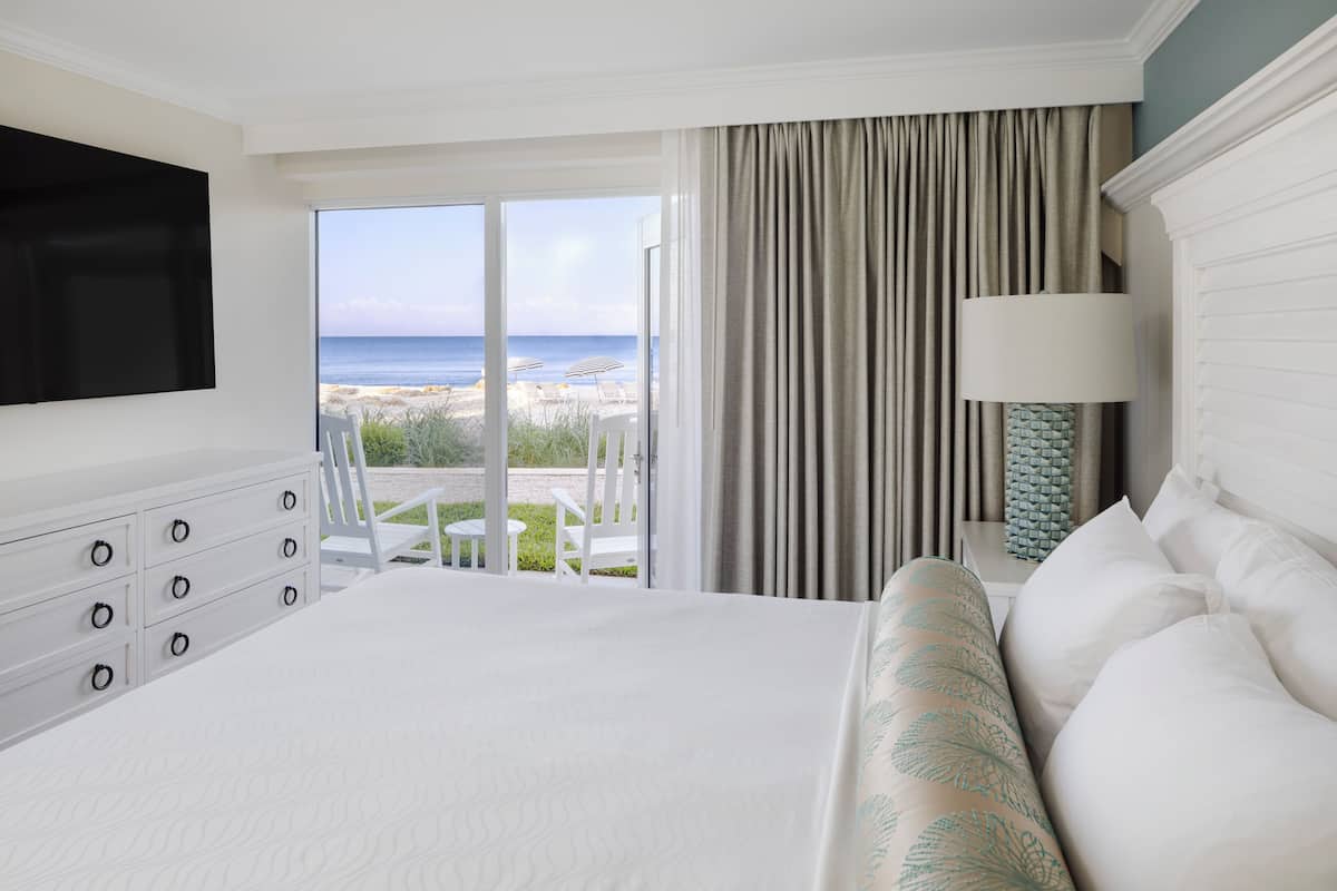 A hotel room with a large bed, a TV, a desk, and a view of the beach.