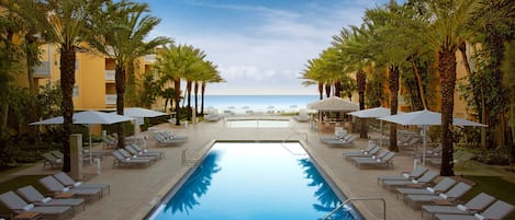 A pool area with lounge chairs and umbrellas, surrounded by palm trees, leading to a beach.