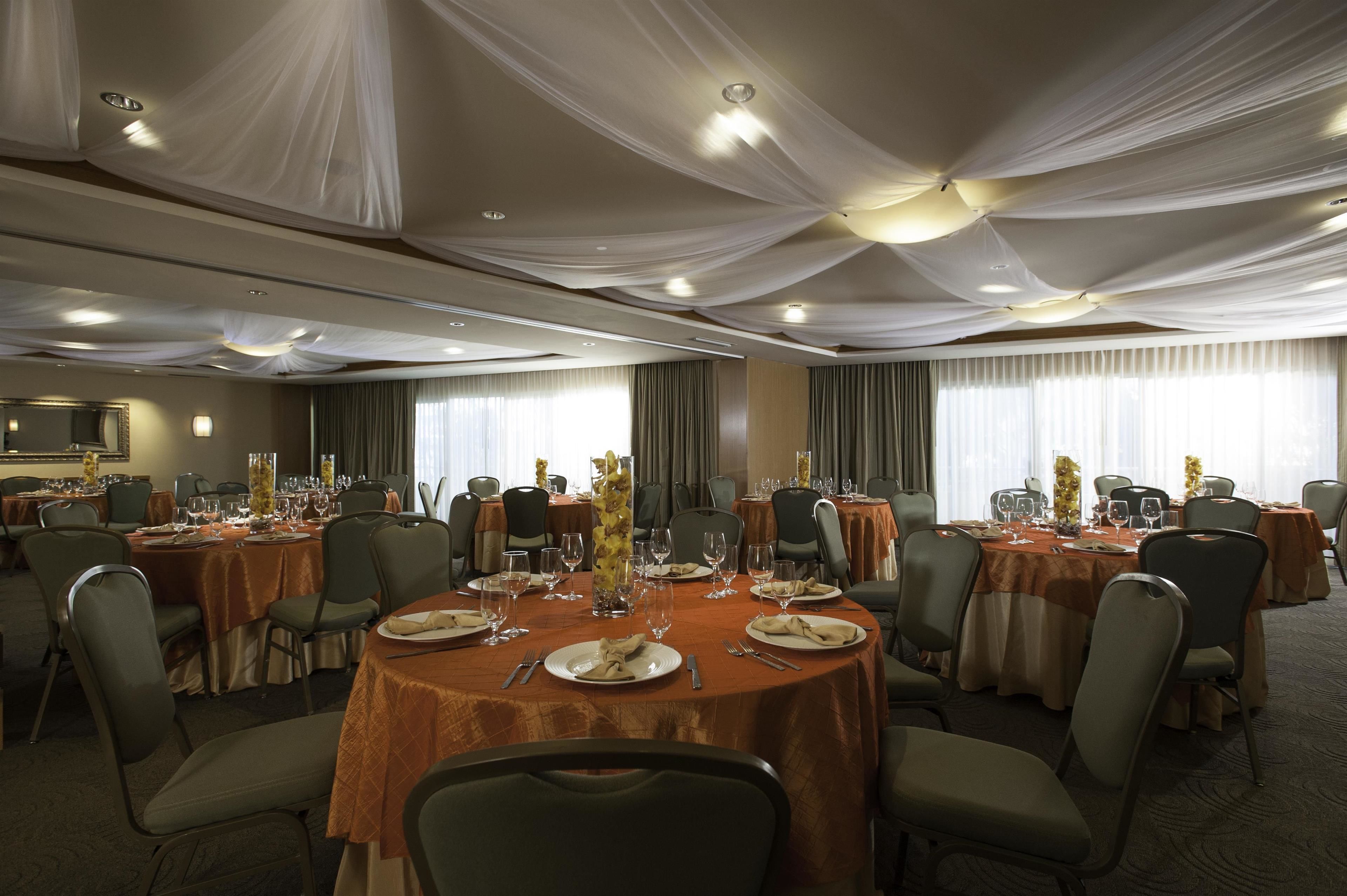 A banquet hall with round tables set for dining, featuring a draped ceiling and large windows with curtains.