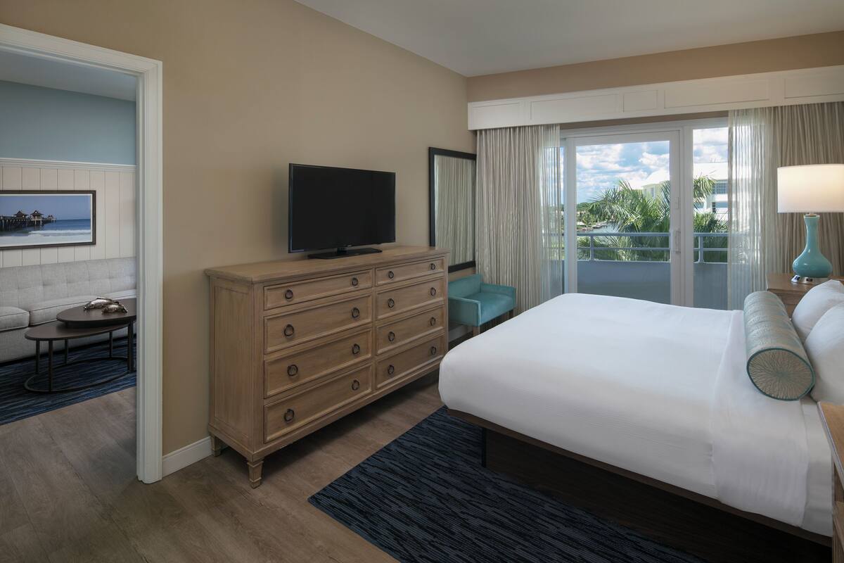 A hotel room with a large bed, a television on a wooden dresser, a balcony with curtains, and a framed picture on the wall.