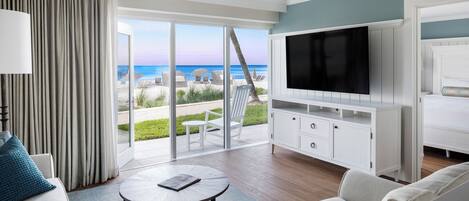 A modern hotel room with a large TV, a sofa, a coffee table, and a balcony with a view of the beach.