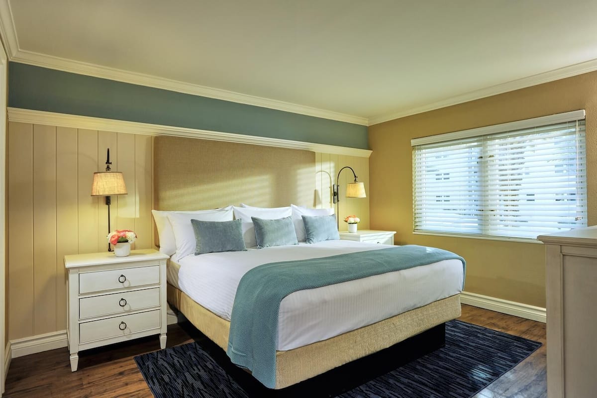 A hotel room with a large bed, two bedside tables, a nightstand, a window with blinds, and a wall-mounted lamp.